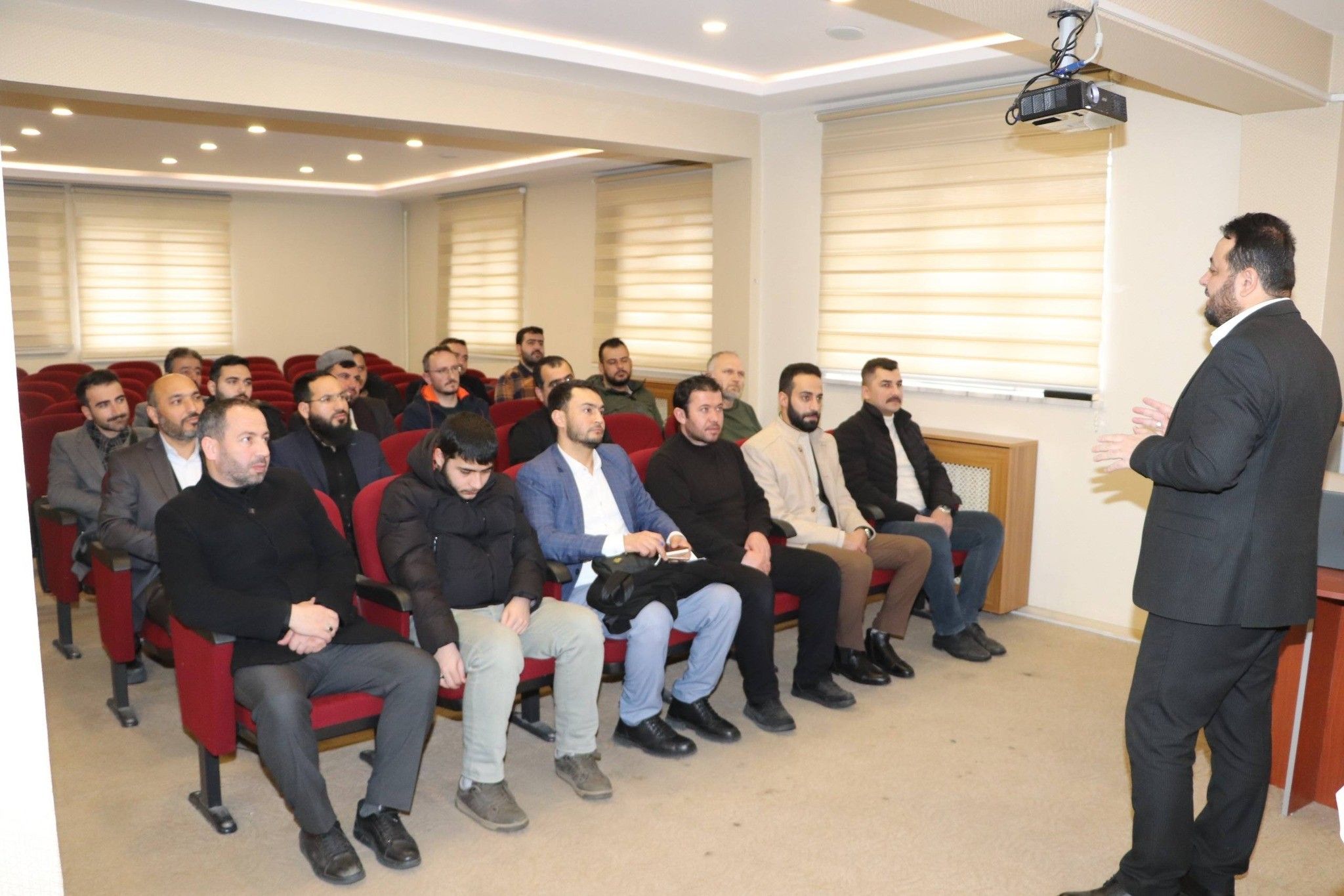 Toplantı salonunda bir kişi oturan erkeklere konuşurken, tavanda projeksiyon cihazı görülüyor.