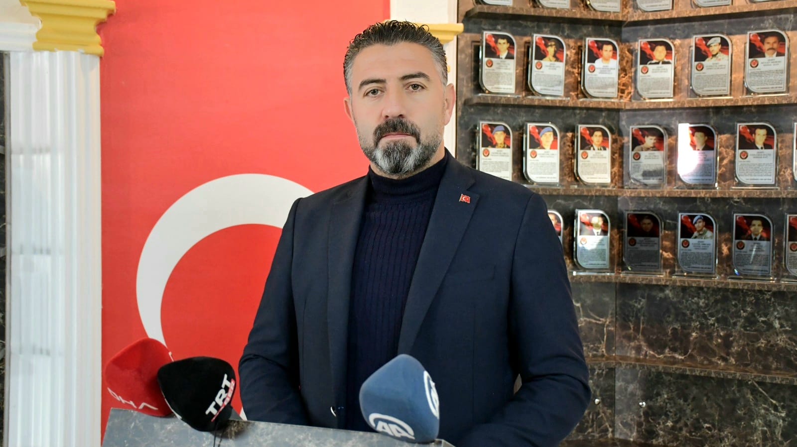 Türk bayrağı arkasında bir kişi, mikrofonların önünde konuşma yapıyor.