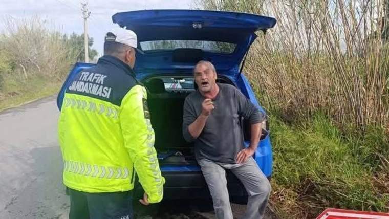 Reflektörlü mont giyen trafik jandarması, açık bagaj kapağı önünde oturan bir adamla konuşuyor.