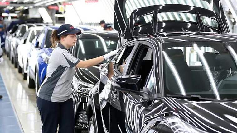 Otomotiv fabrikasında iş kıyafetli bir kadın, üretim hattındaki siyah otomobilin kapısını kontrol ediyor.