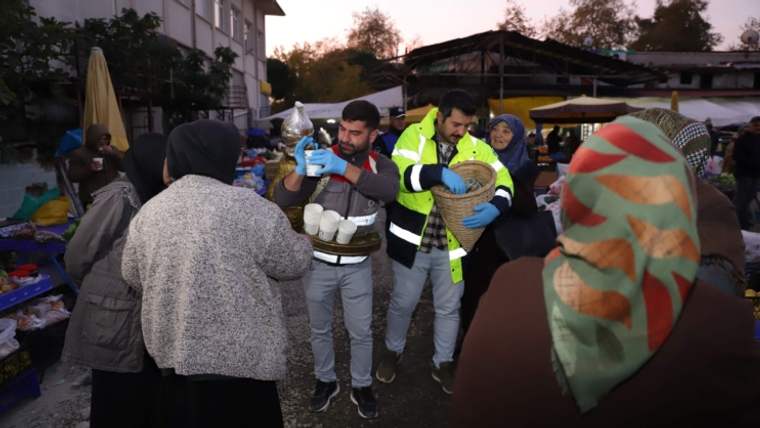 Pazar alanında, elinde yiyecek ve içecek taşıyan iki kişi, çevresindeki esnafa yardım ediyor.