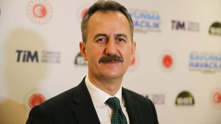 Takım elbiseli, bıyıklı bir erkek, üzerinde çeşitli kurum logoları bulunan pano önünde duruyor.