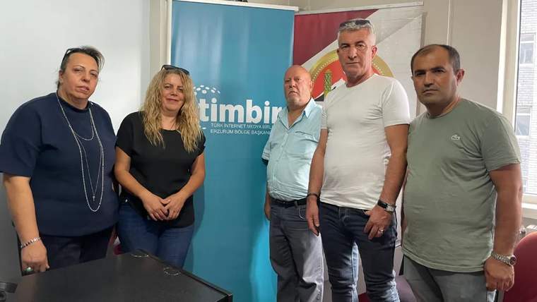 İç mekânda, üzerinde timbir yazısı bulunan afişin önünde duran dört erkek ve iki kadın