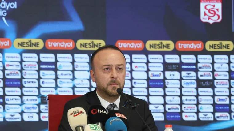Sponsor logolarının yer aldığı pano önünde bir kişi, çok sayıda mikrofonun bulunduğu masada konuşuyor.