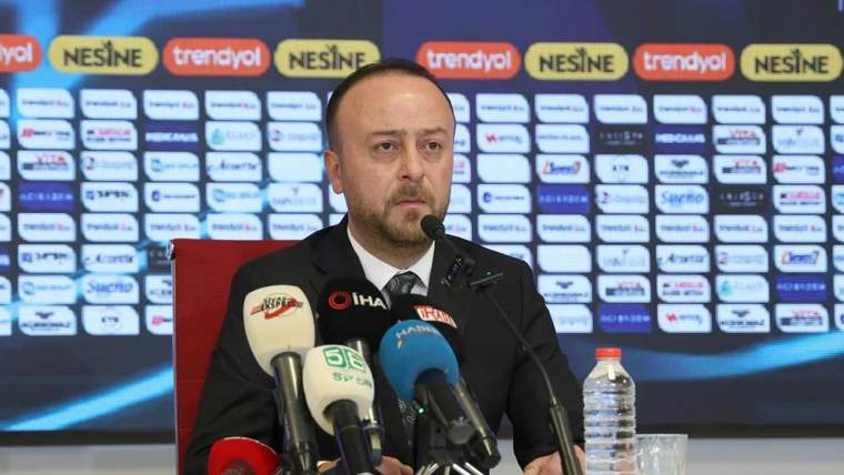 Sponsor logolarının yer aldığı pano önünde bir kişi, çok sayıda mikrofonun bulunduğu masada konuşuyor.