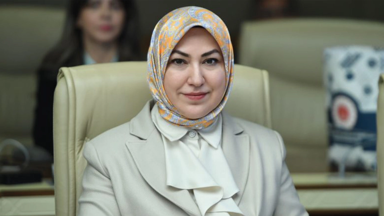Desenli başörtülü bir kadın, açık renk ceketle meclis salonunu andıran bir ortamda koltukta oturuyor.