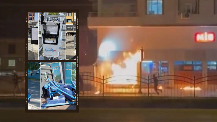 Alevler içinde bir işyeri, önünde bir kişi koşuyor; yan tarafta devrilmiş mavi malzemeler ve bir ATM görüntüsü var.