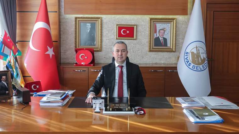 Sivas Belediye Başkanı Adem Uzun, makamında oturuyor, arkasında Türk bayrağı ve Sivas Belediyesi logosu var.