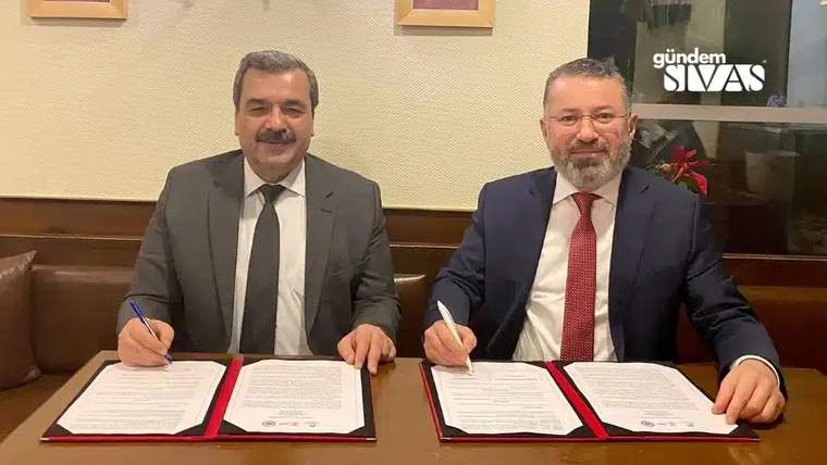 Masada oturan iki kişi, önlerindeki belgeleri kalemle imzalarken kameraya bakıyor.