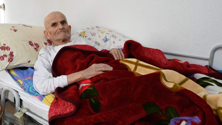Yatakta yatan yaşlı bir adam, üzerine kırmızı bir battaniye örtülü.