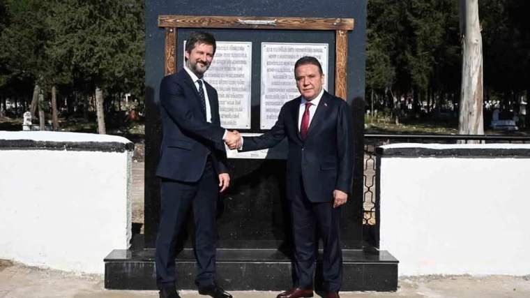 Takım elbiseli iki kişi, yazılı bir anıt levhasının önünde el sıkışırken görülüyor.
