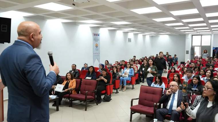Konferans salonunda mikrofonla konuşan bir kişi, karşısında oturan kalabalık dinleyici grubuna sesleniyor.