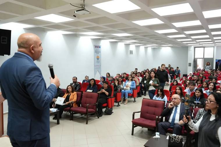Konferans salonunda mikrofonla konuşan bir kişi, karşısında oturan kalabalık dinleyici grubuna sesleniyor.