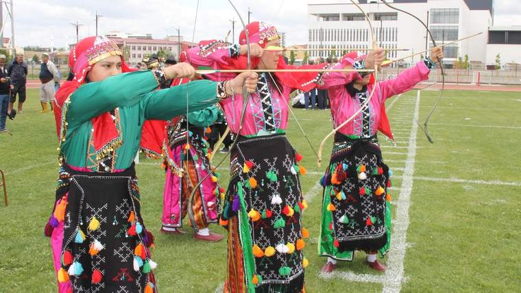 Geleneksel kıyafetler giyen dört sporcu, çim sahada yaylarını aynı hedefe doğru geriyor.