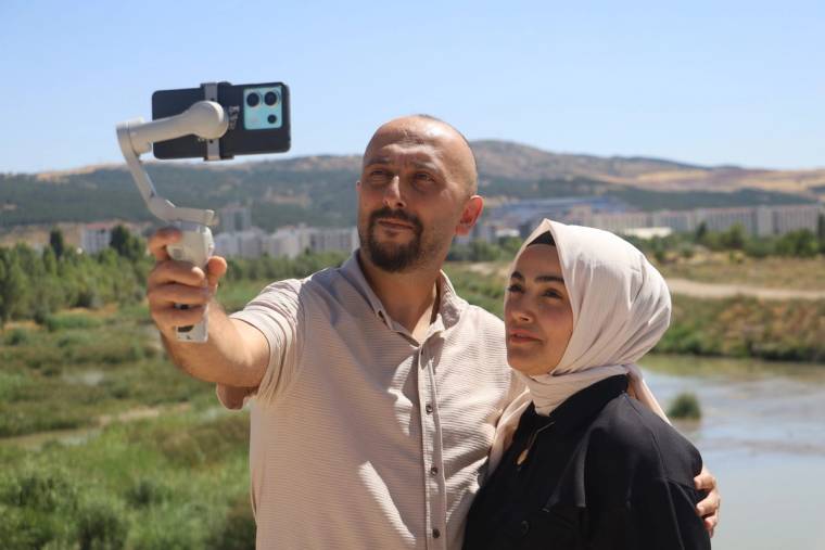 Bir erkek ve başörtülü bir kadın, nehir ve tepeler önünde telefonla özçekim yapıyor.
