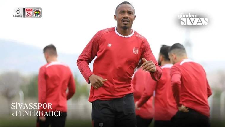Kırmızı antrenman üstü ve siyah şort giyen bir futbolcu önde koşarken, arka planda takım arkadaşları görülüyor.