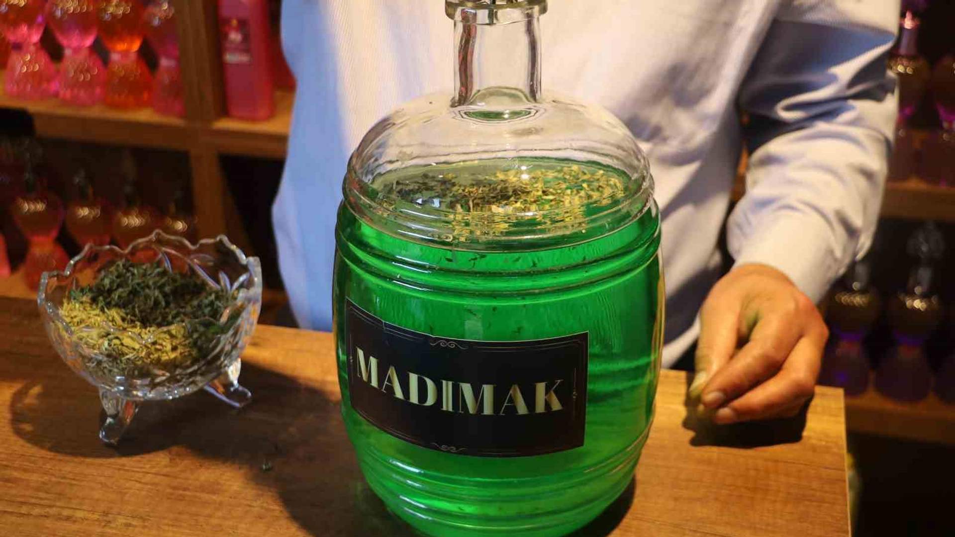 Üzerinde “MADIMAK” yazan yeşil cam kavanoz, tezgâhta otlarla dolu bir kasenin yanında duruyor.
