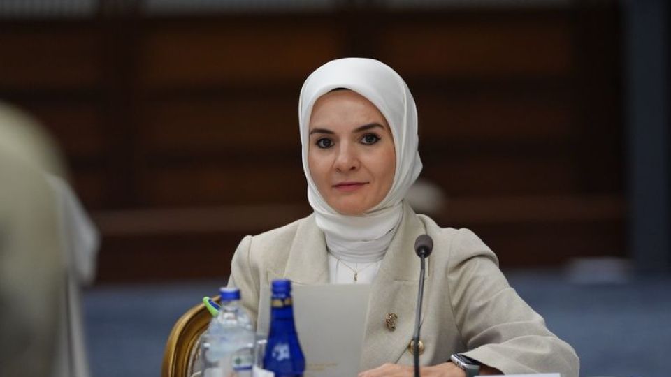 Başörtülü bir kadın, mikrofon ve dizüstü bilgisayarın bulunduğu toplantı masasında oturuyor.