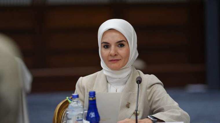 Başörtülü bir kadın, mikrofon ve dizüstü bilgisayarın bulunduğu toplantı masasında oturuyor.