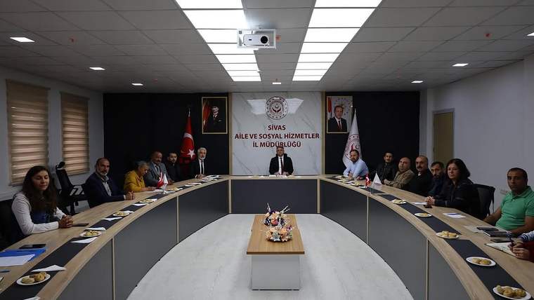 Toplantı salonunda uzun masanın çevresinde oturan kişiler, duvardaki kurum panosu önünde görüşme yapıyor.