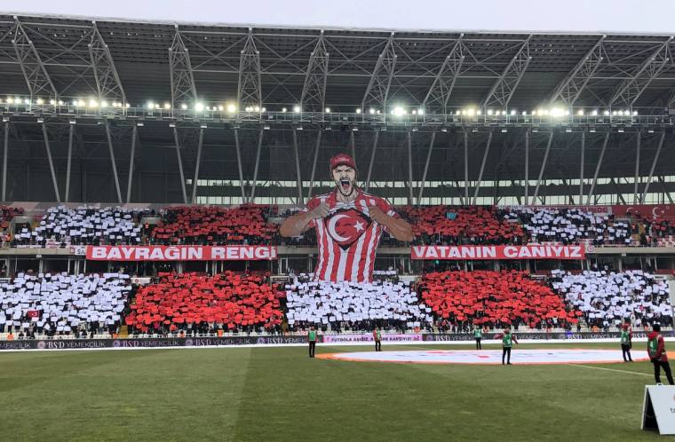Tribünlerde kırmızı beyaz kartonlarla Türk bayrağı ve büyük bir futbolcu figürü oluşturuluyor.