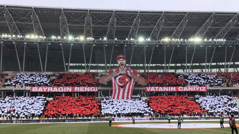 Tribünlerde kırmızı beyaz kartonlarla Türk bayrağı ve büyük bir futbolcu figürü oluşturuluyor.