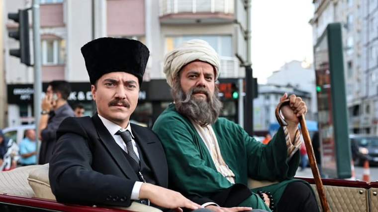 Açık araçta oturan, dönem kostümü giymiş iki sakallı ve bıyıklı erkek şehir caddesinde görülüyor.