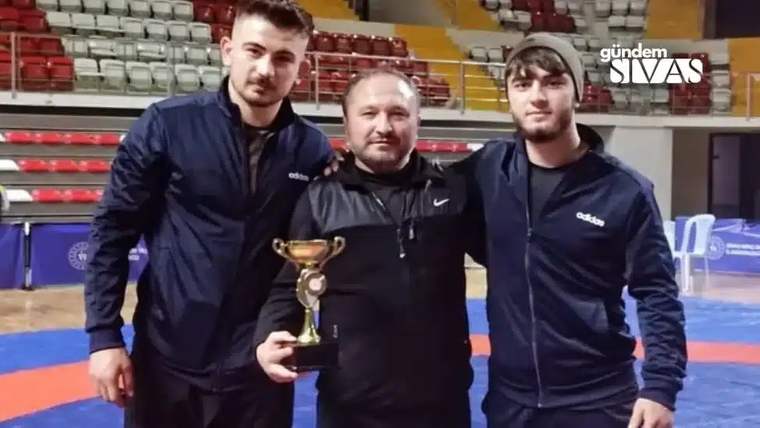 Şampiyonluk kupası tutan bir antrenör ve iki sporcu, spor salonunda poz veriyor.
