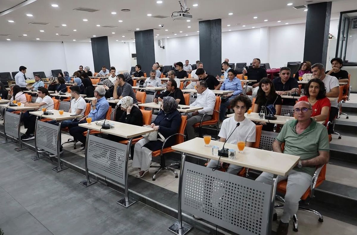 Sıralı masalarda oturan kalabalık bir grup, önlerindeki mikrofon ve turuncu içeceklerle toplantı salonunda görülüyor.