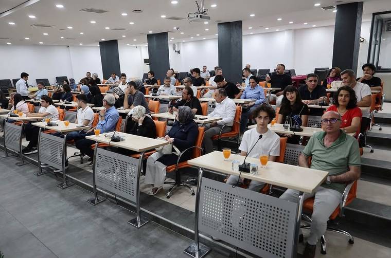 Sıralı masalarda oturan kalabalık bir grup, önlerindeki mikrofon ve turuncu içeceklerle toplantı salonunda görülüyor.