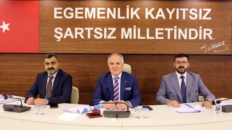 Ahşap panonun önünde, masada yan yana oturan üç takım elbiseli kişi görülüyor.