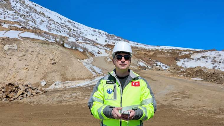 Beyaz kask takan bir kişi, taş ocağında duruyor; arka planda toprak ve kayalık alanlar var.
