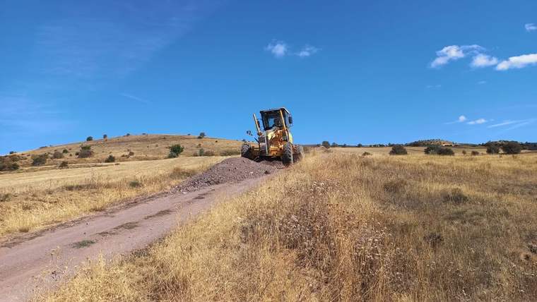 Kuru otlarla kaplı arazide bir iş makinesi, taşlı toprak yol üzerinde ilerlerken görülüyor.