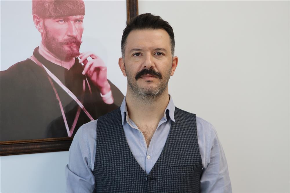 Bir adam, gömlek ve yelek giymiş, arkasında bir çerçeveli portre var.