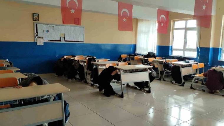 Türk bayrakları asılı sınıfta öğrenciler sıraların altına çömelmiş, bazı sıralar boş duruyor.
