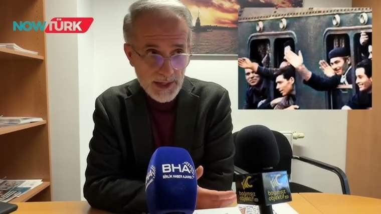 Bir masanın arkasında oturan bir kişi, mikrofonların önünde konuşuyor.