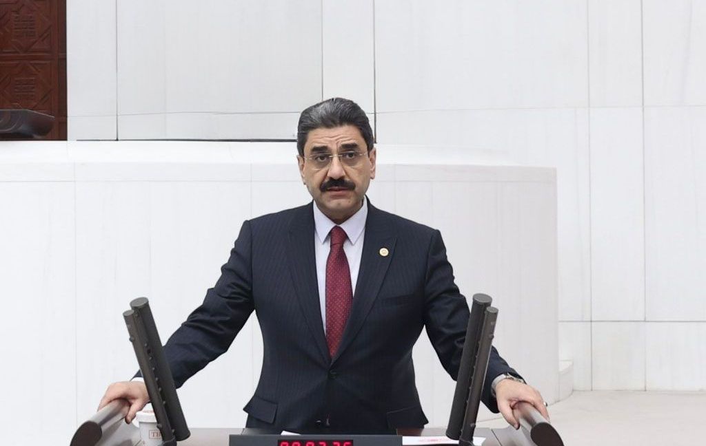 Kürsüde konuşan bir kişi, arka planda beyaz bir duvar ve masa var.