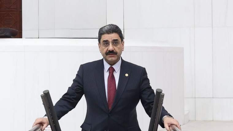 Kürsüde konuşan bir kişi, arka planda beyaz bir duvar ve masa var.