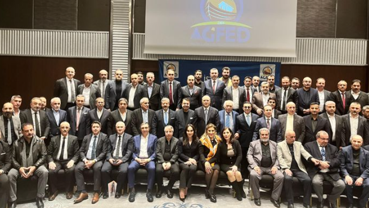 Bir grup insan, bir etkinlikte poz veriyor; arka planda AGHED logosu ve mavi bir pankart var.