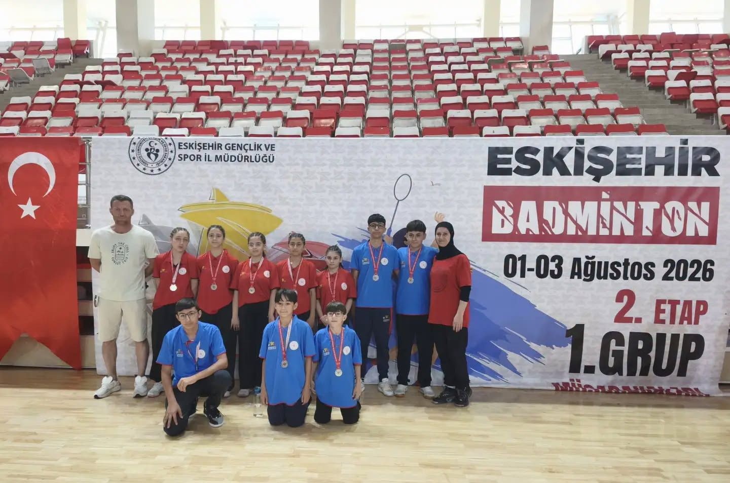 Badminton takımı, madalyalarıyla birlikte bir arada poz veriyor. Arkada büyük bir pankart var.