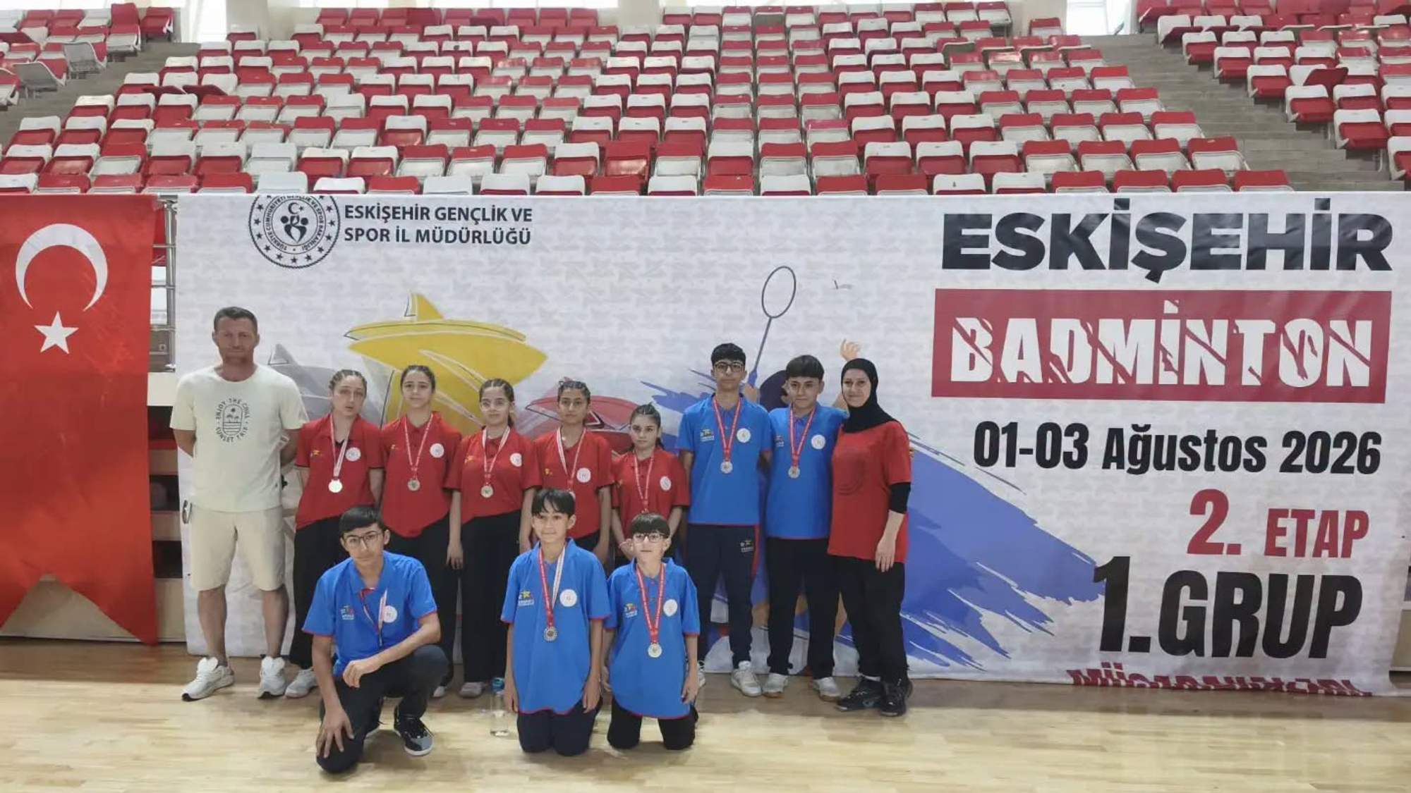 Badminton takımı, madalyalarıyla birlikte bir arada poz veriyor. Arkada büyük bir pankart var.
