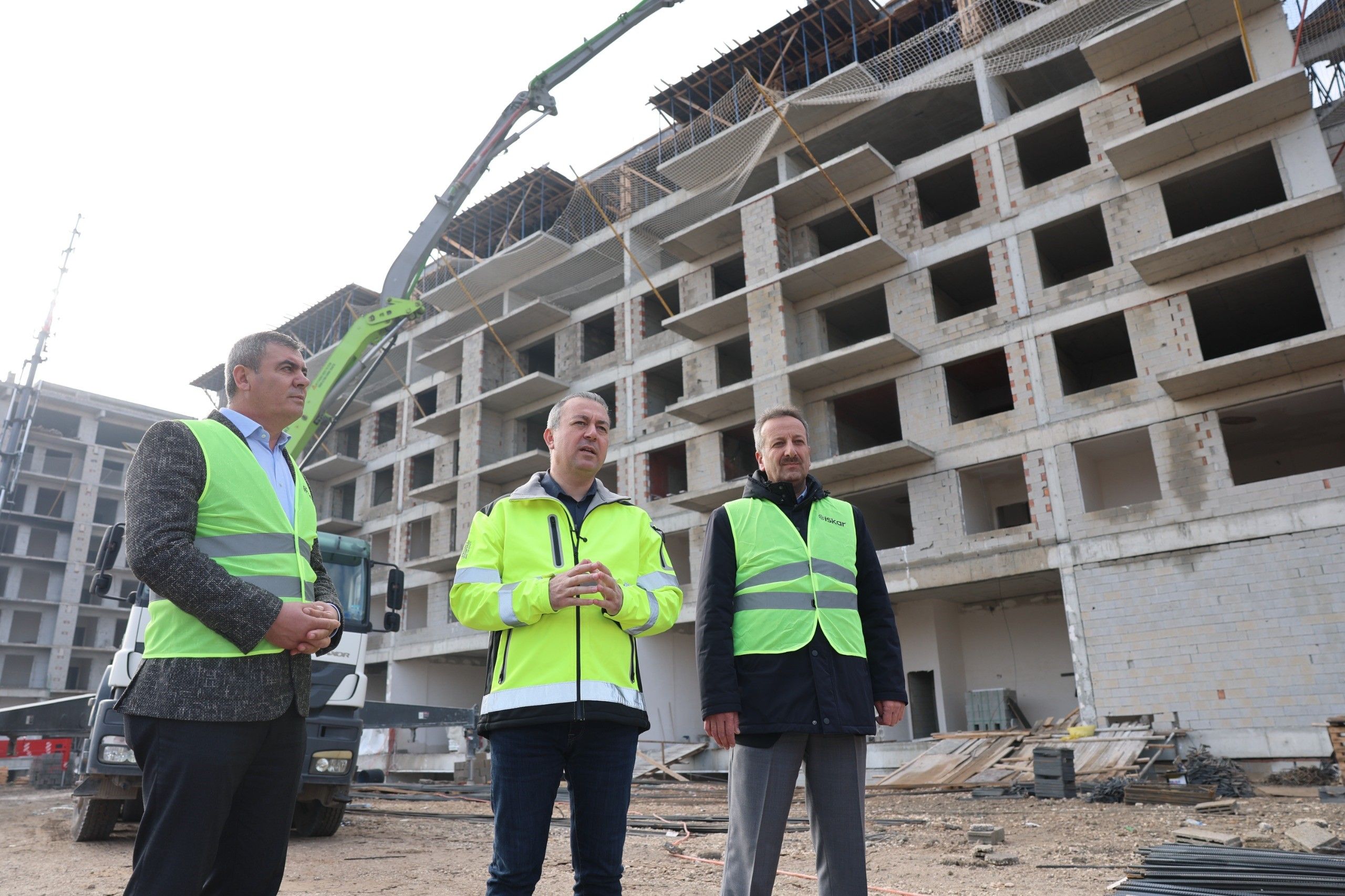 İnşaat alanında üç kişi, tamamlanmamış çok katlı betonarme binanın önünde duruyor.