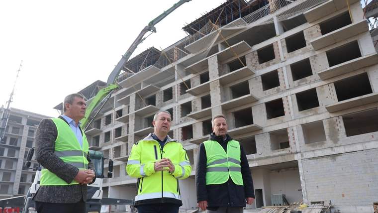 İnşaat alanında üç kişi, tamamlanmamış çok katlı betonarme binanın önünde duruyor.