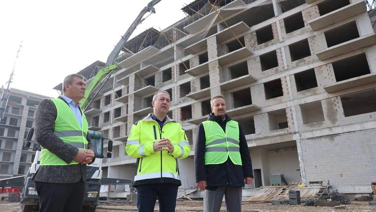 İnşaat alanında üç kişi, tamamlanmamış çok katlı betonarme binanın önünde duruyor.
