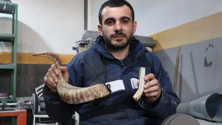 Bir kişi, elinde bir bıçak sapı ve bir boynuz parçası tutuyor, arka planda atölye ekipmanları görünüyor.