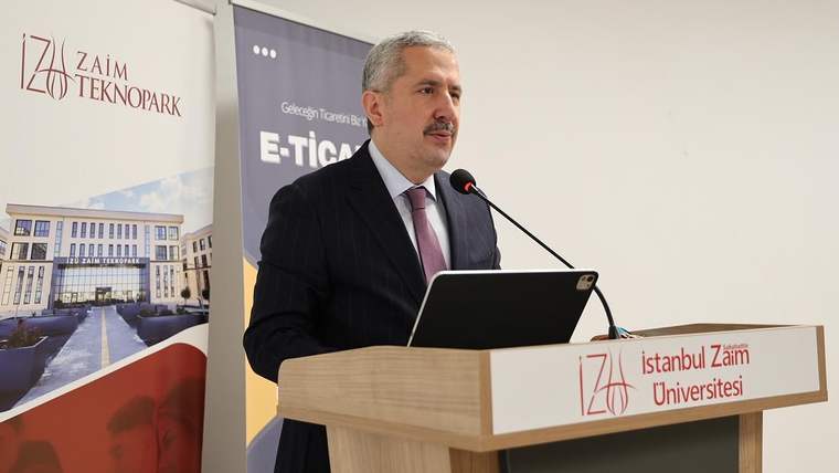 Bir kişi, podiumda mikrofonla konuşuyor; arka planda üniversite logosu ve e-ticaret afişi var.