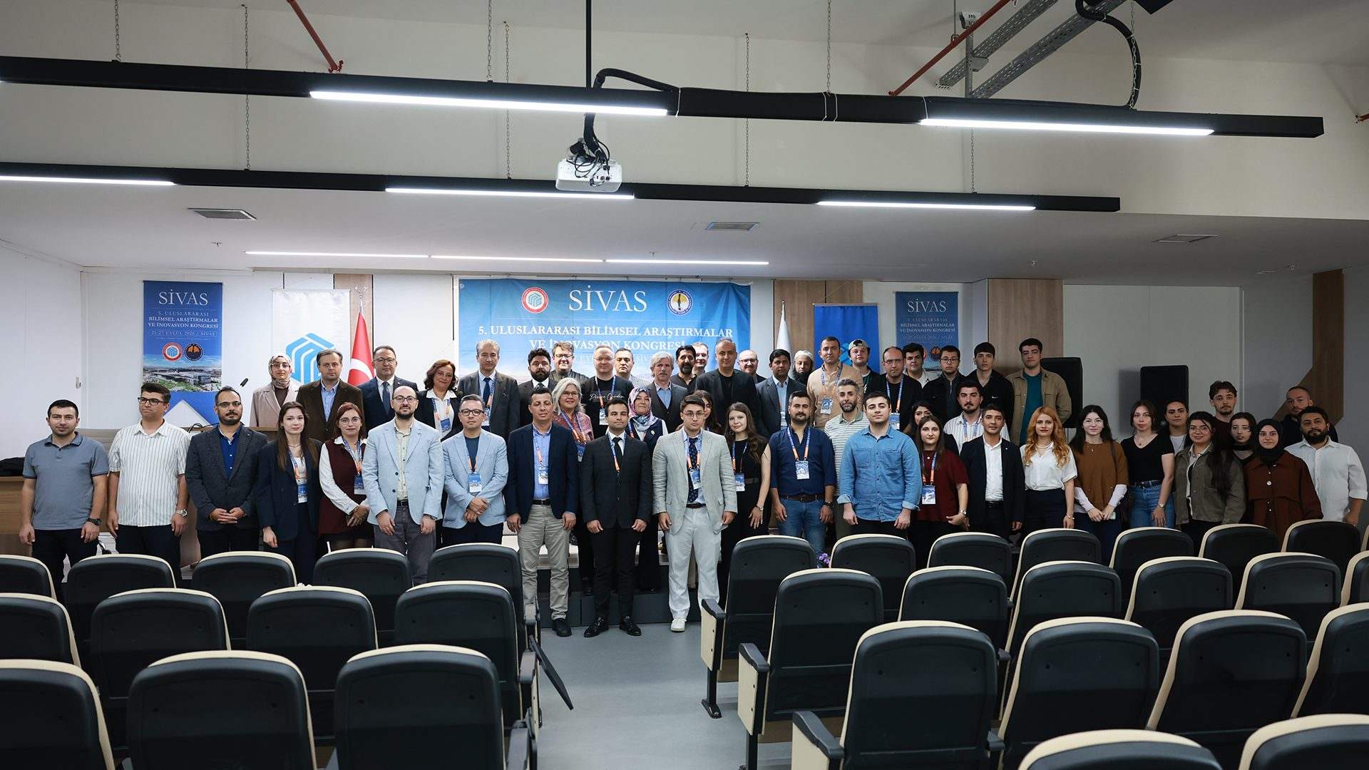 Konferans salonunda, arkalarında Sivas yazılı afişler bulunan kalabalık bir grup birlikte poz veriyor.