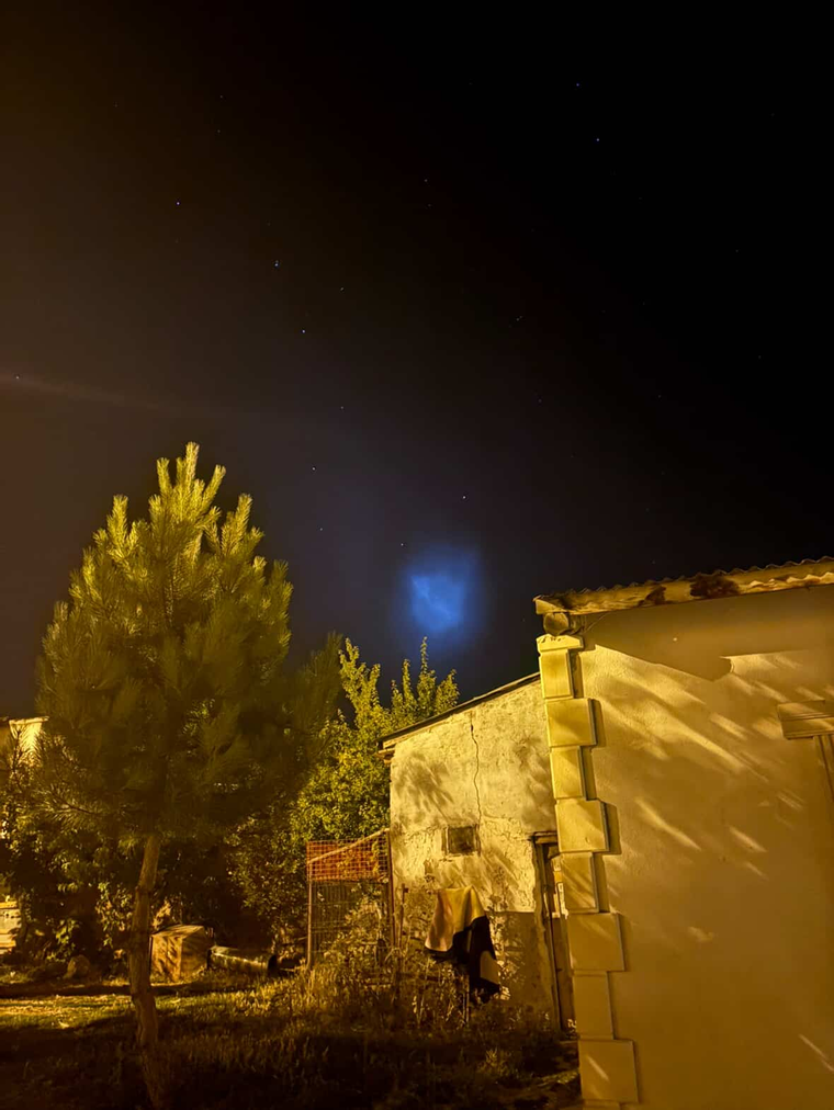 Gece gökyüzünde ağaçların ve evin üzerinde mavi ışık saçan belirsiz bir cisim görülüyor.