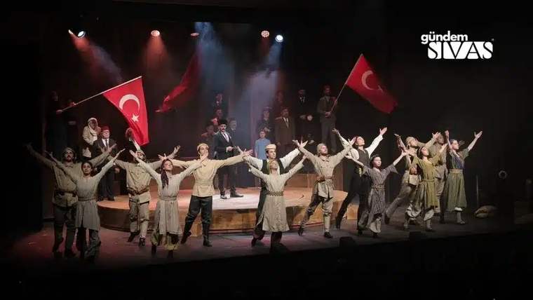Tiyatro sahnesinde çok sayıda oyuncu, Türk bayrakları ve renkli sahne ışıklarıyla birlikte selam veriyor.