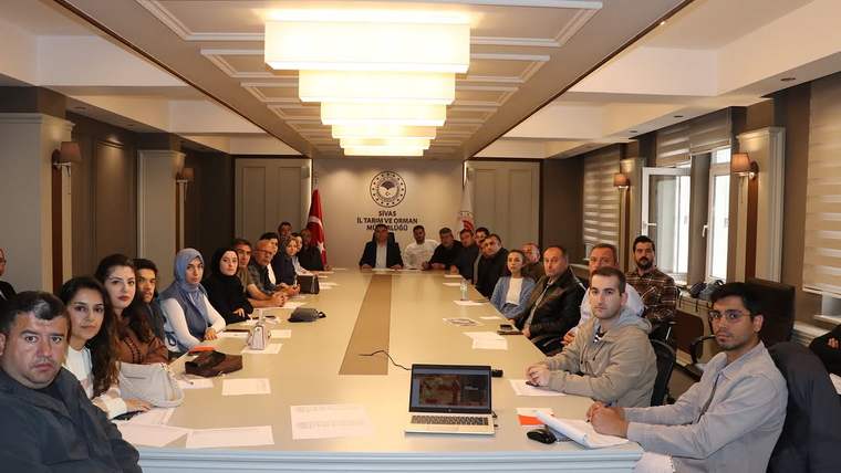 Toplantı salonunda uzun masa çevresinde oturan kalabalık grup, Türk bayrağı ve duvar amblemi görülüyor.